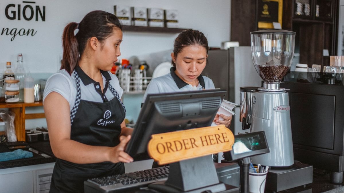 Barista di balik meja kasir kedai kopi dengan mesin kasir dan peralatan kopi
