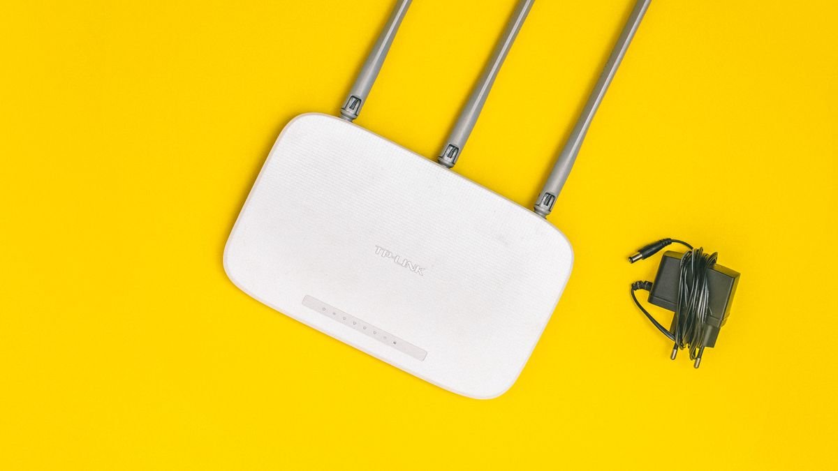 Router Wi-Fi putih dengan tiga antena dan kabel di atas latar kuning